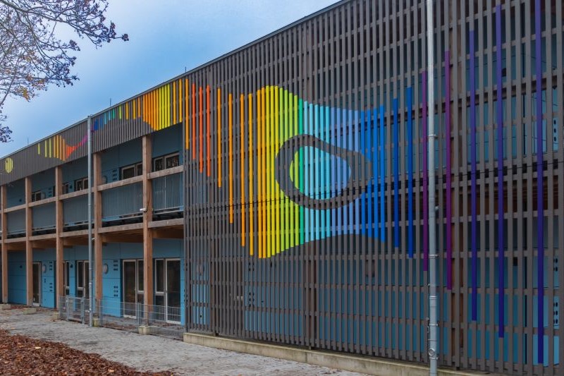 Blick von rechts auf die graue Fassade in holzbauweise mit dem farbigen Kunstwerk auf den Latten